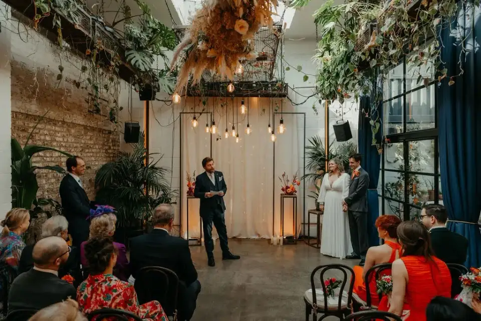 Couple standing before the celebrant under hanging Edison bulbs and dried flowers as guests look on