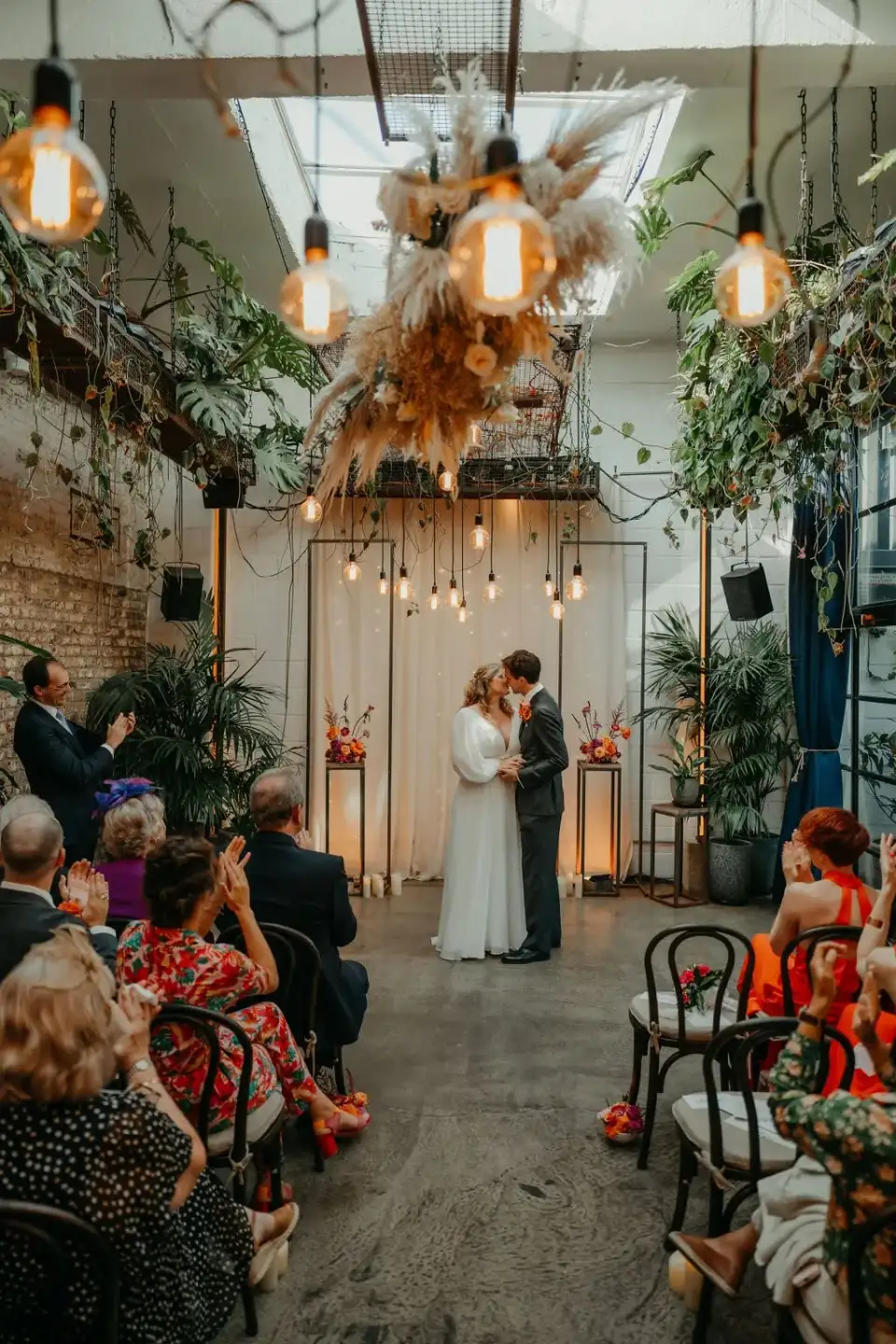 Couple sharing a kiss at the end of the aisle under a dried flower installation and hanging bulbs