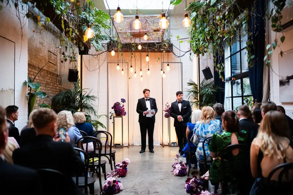 Groom and best man waiting at the front of the Winter Garden aisle, lined with deep pink flowers