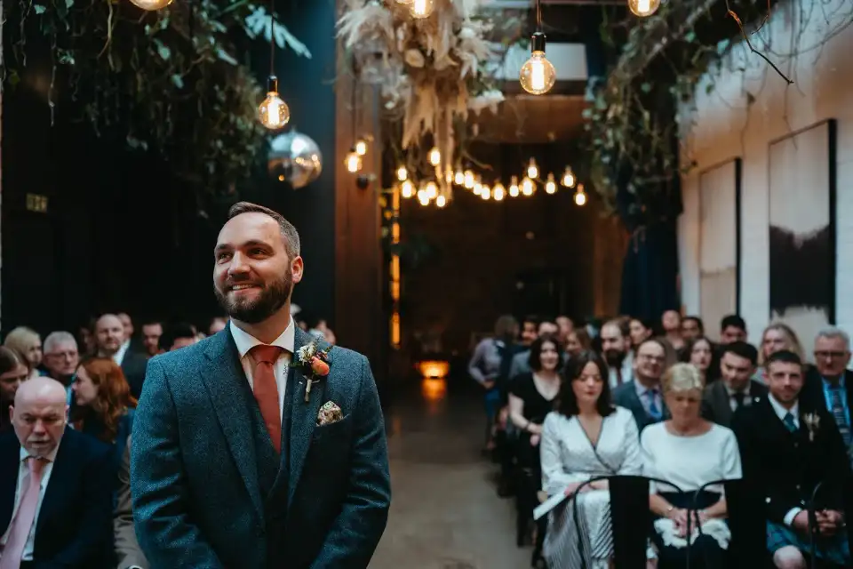 Groom smiling at the front of the aisle as seated guests wait under festoon bulbs and a disco ball