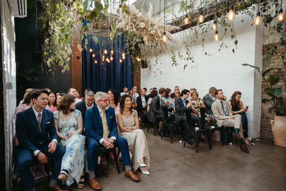 Seated wedding guests smiling towards the aisle beneath trailing greenery and navy curtains