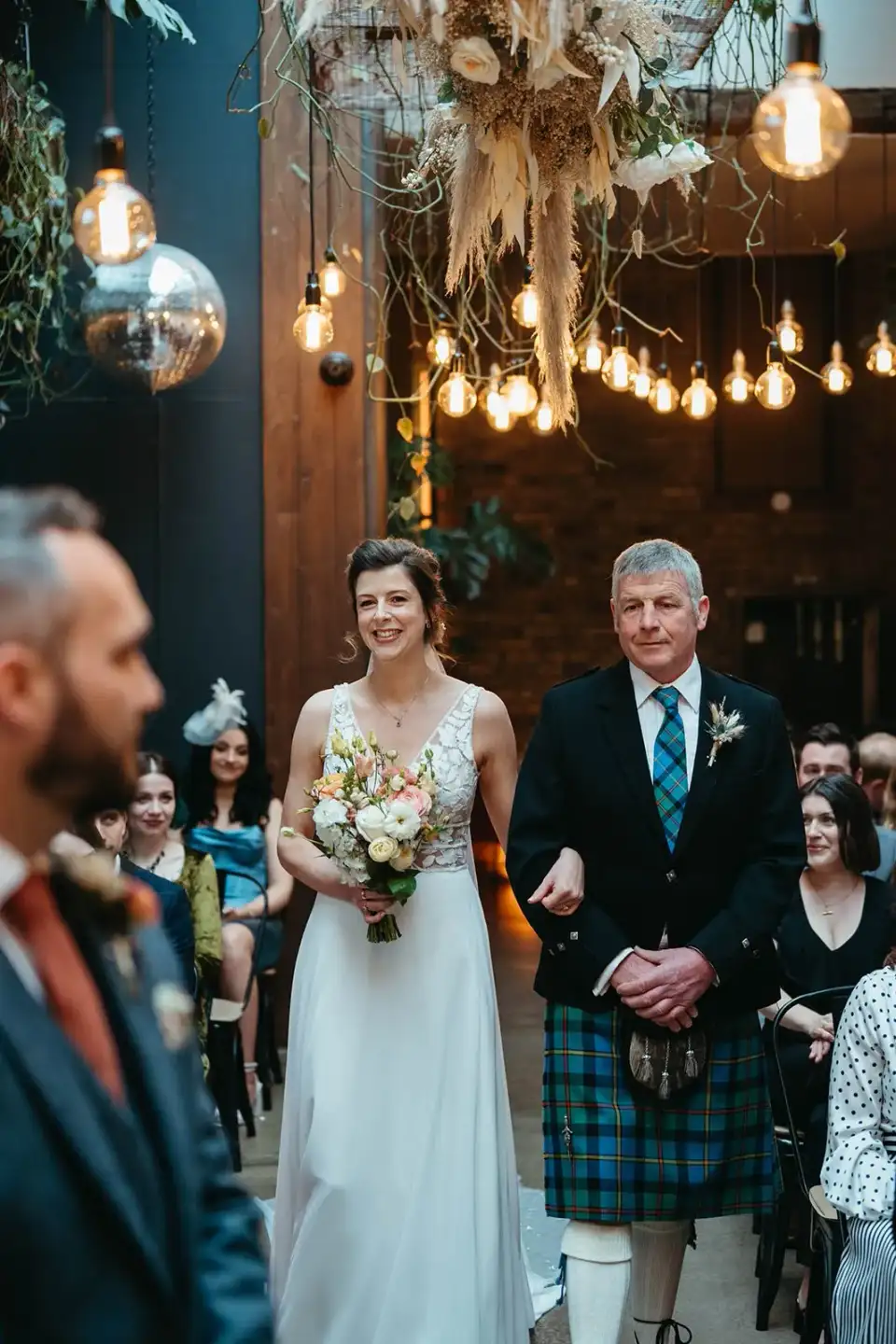 Bride walking down the aisle with her father in a kilt, guests turning to watch under festoon lights