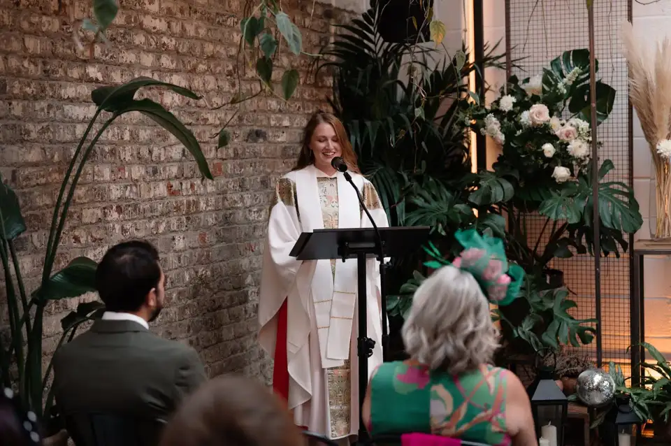 Celebrant in white robes smiling at the lectern beside potted palms and exposed brick