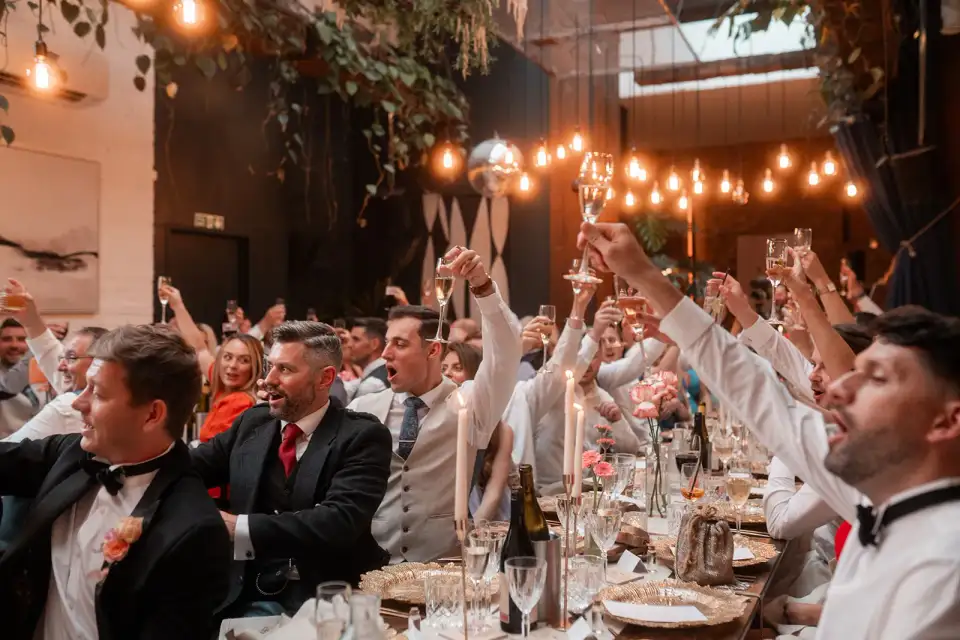 Guests cheering with raised glasses across candlelit tables under strings of Edison bulbs