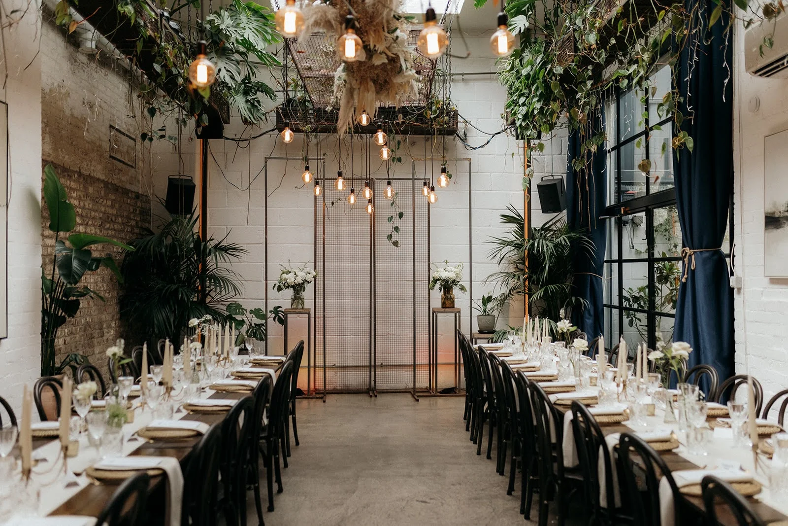 The Hackney's main hall set for a wedding - long banquet tables with bentwood chairs flanking the central aisle, hanging Edison bulbs and pampas, white brick walls, deep navy curtains on factory windows. Photo by Caitlin and Jones.