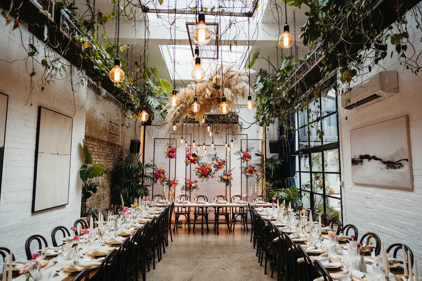 The Winter Garden at The Hackney laid for dinner: two long tables with candles, hanging plants and bare bulbs, and bright flowers on mesh screens at the far end